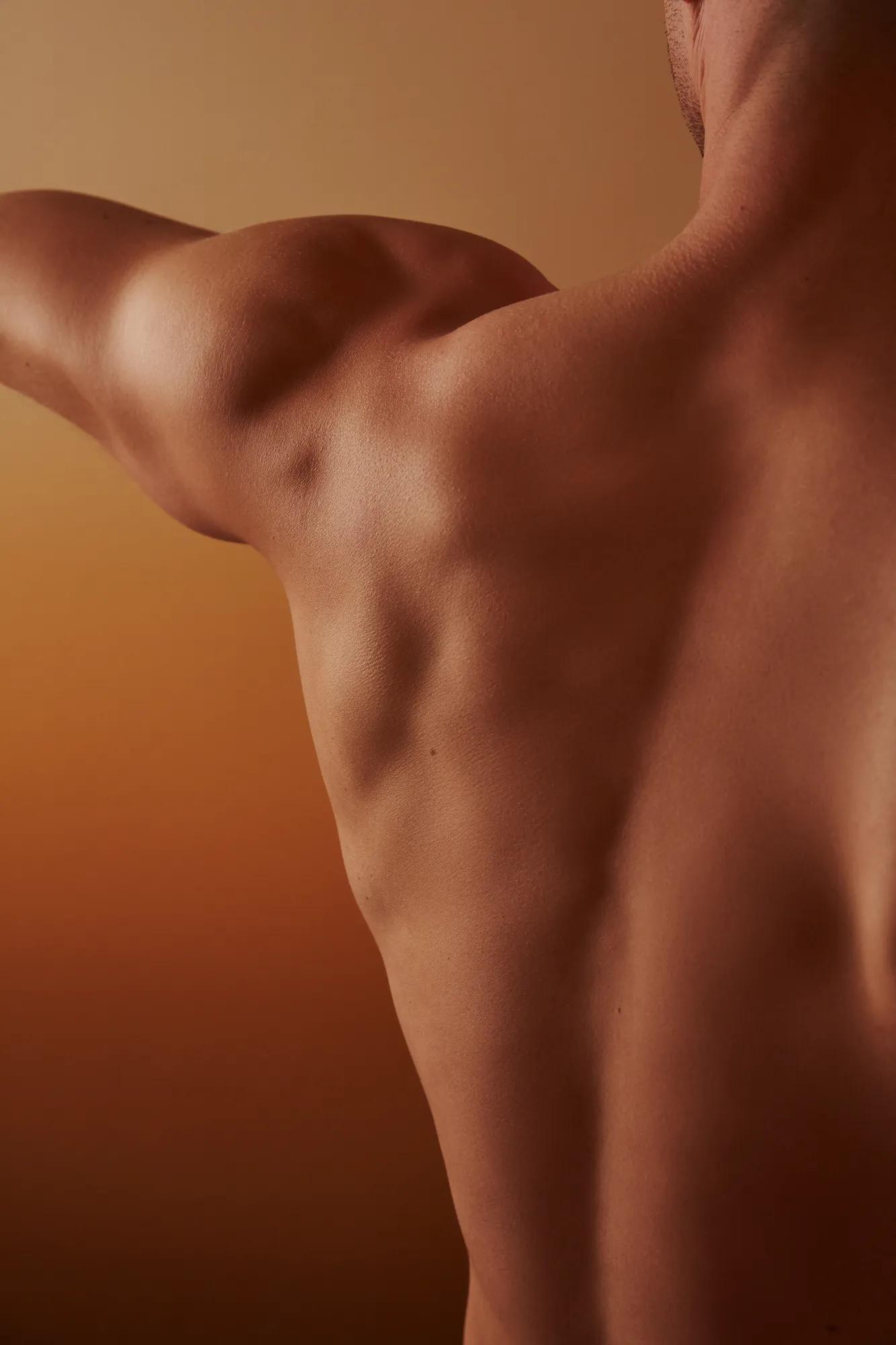 Close-up of a man’s bare back and shoulder with arm extended, lit in warm amber tones