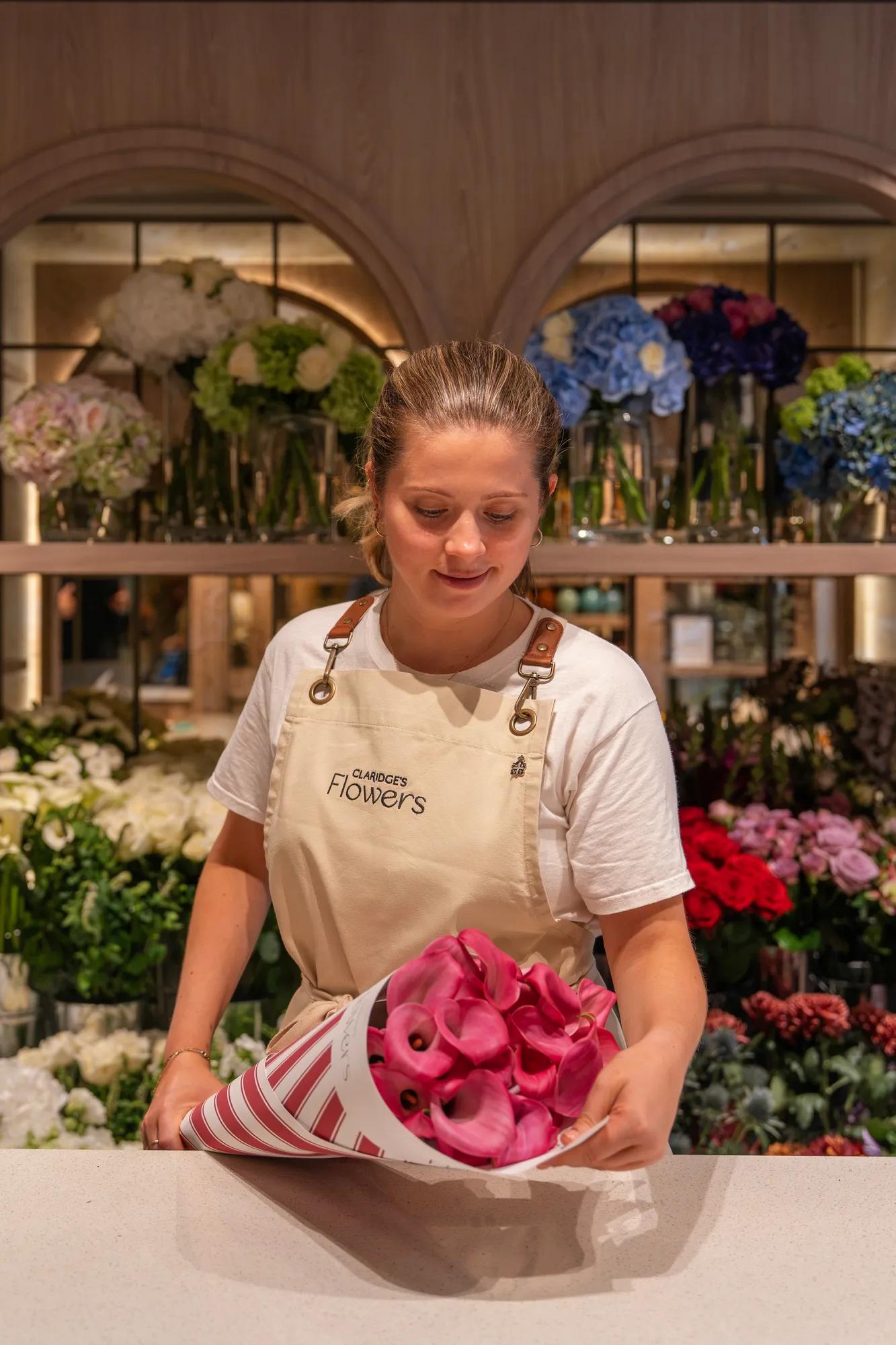 Florist wrapping a bouquet of pink calla lilies at a flower counter, surrounded by colourful fresh blooms.
