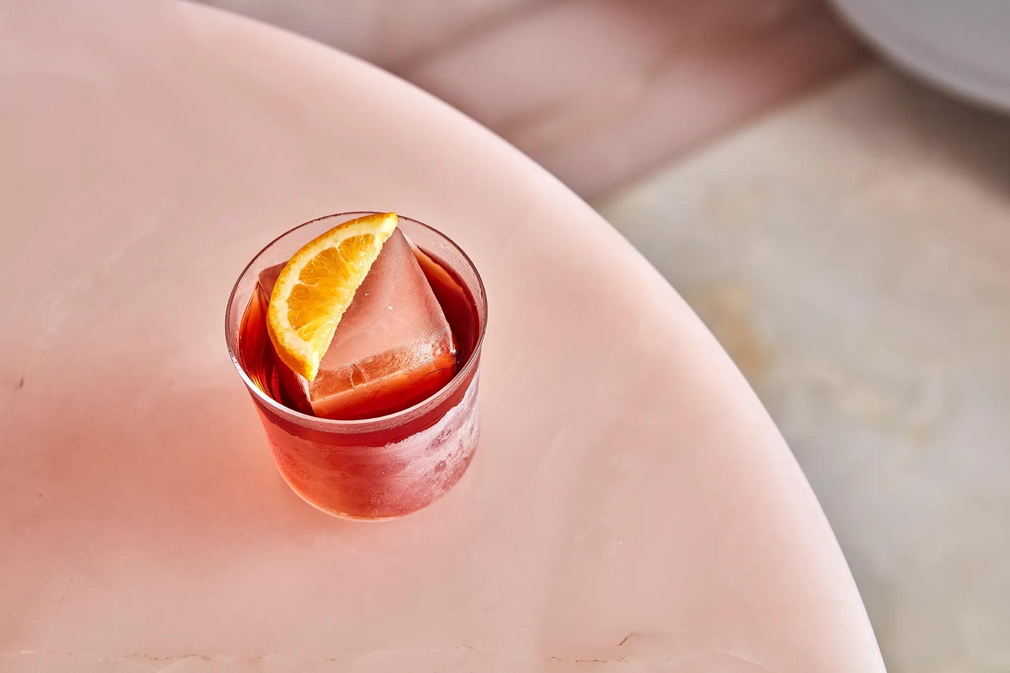 Red cocktail with a large ice cube and orange wedge garnish on a pale pink marble table.