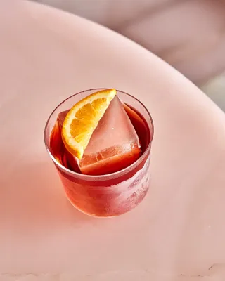Red cocktail with a large ice cube and orange wedge garnish on a pale pink marble table.