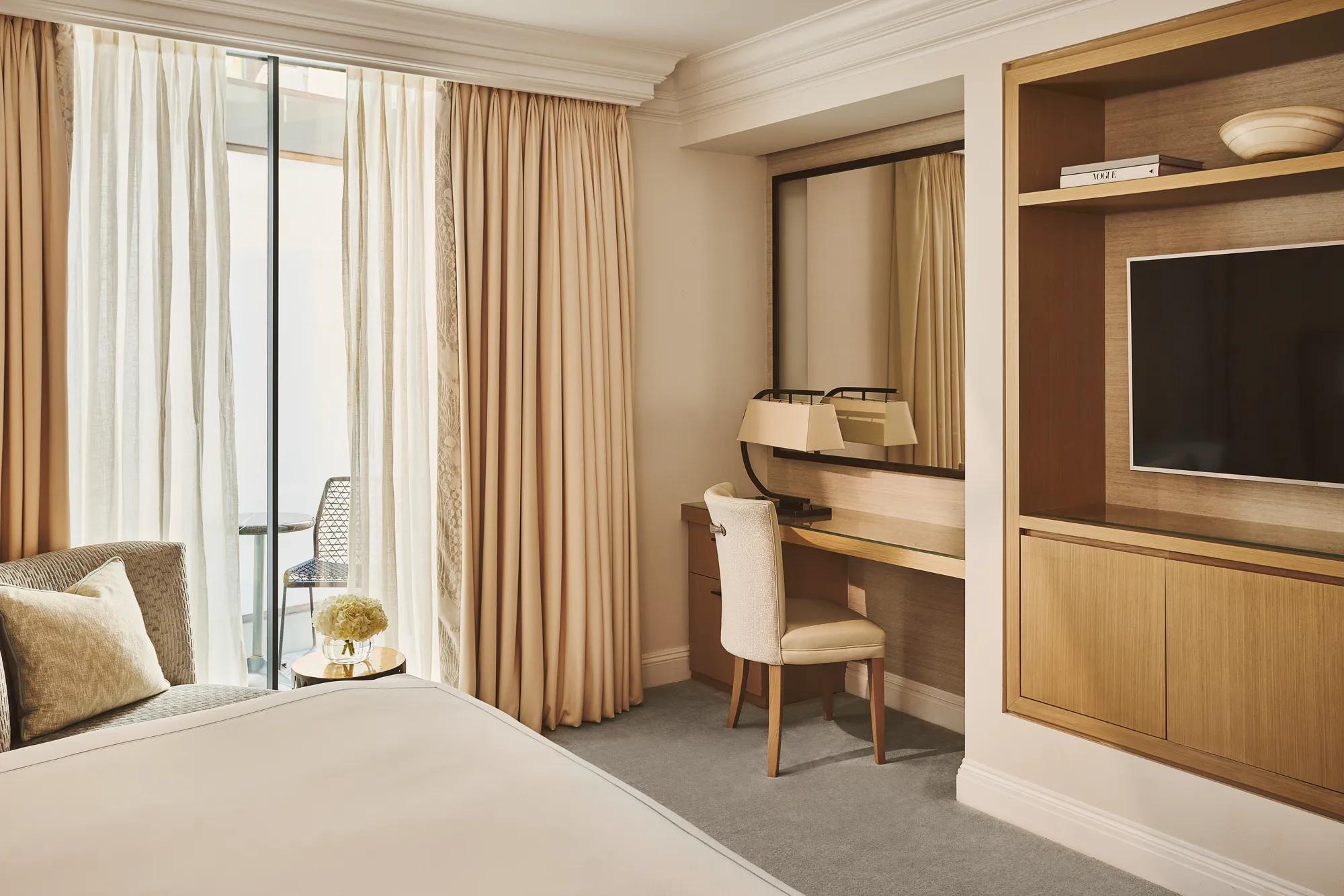 Bedroom with a bed, upholstered chair and desk beside floor-to-ceiling curtains, with a wall-mounted television and shelving unit.