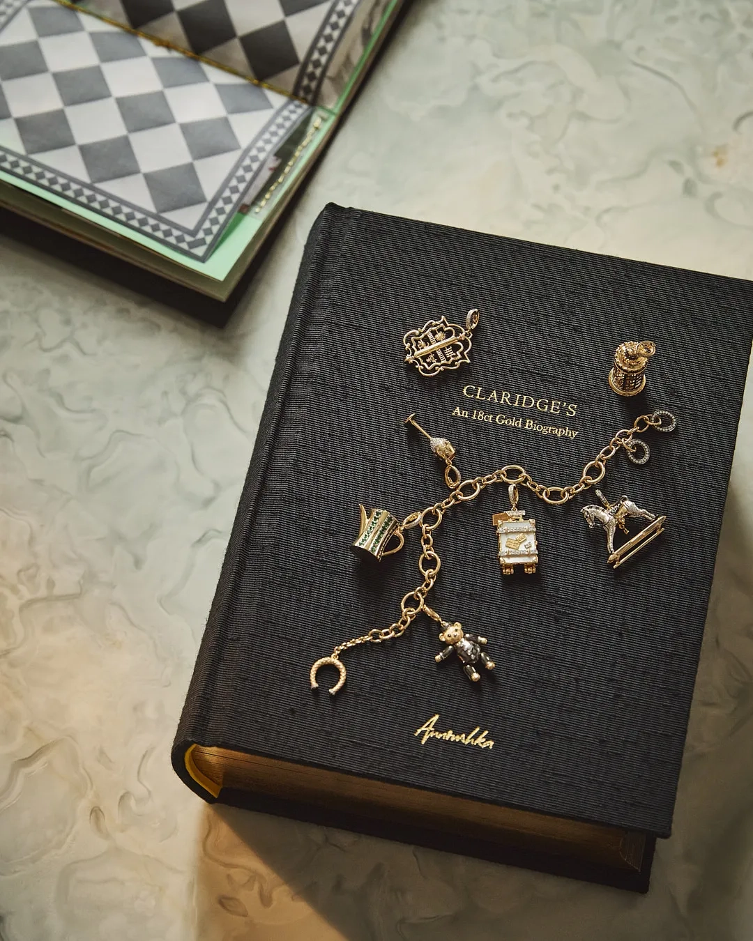 A black book titled Claridge’s: An 18ct Gold Biography lies on a marble surface, adorned with a gold charm bracelet and several individual gold charms shaped like a crown, a teddy bear, and miniature hotel motifs.