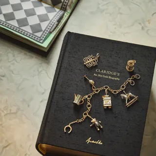 A black book titled Claridge’s: An 18ct Gold Biography lies on a marble surface, adorned with a gold charm bracelet and several individual gold charms shaped like a crown, a teddy bear, and miniature hotel motifs.