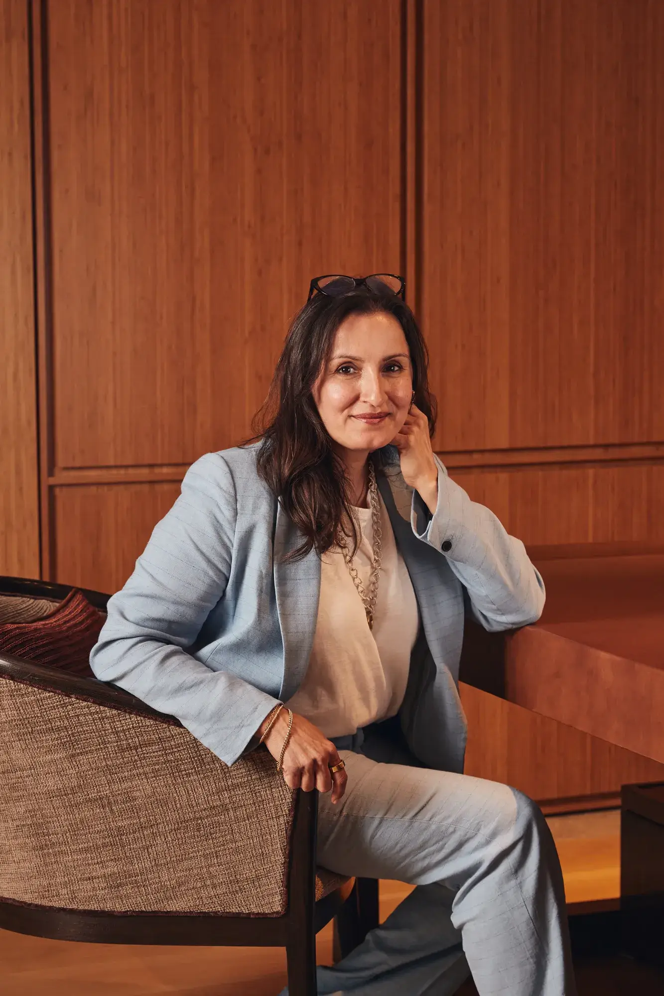 Person seated in an upholstered chair, wearing a light blue suit, resting their head on one hand in a wood-panelled interior.