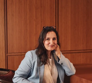 Person seated in an upholstered chair, wearing a light blue suit, resting their head on one hand in a wood-panelled interior.