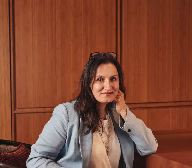 Person seated in an upholstered chair, wearing a light blue suit, resting their head on one hand in a wood-panelled interior.
