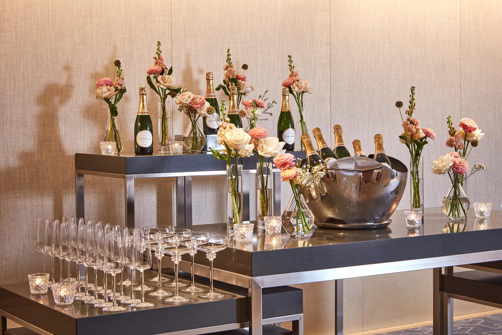 Champagne bar setup with bottles on ice, flutes, and vases of pastel flowers on modern black tables.