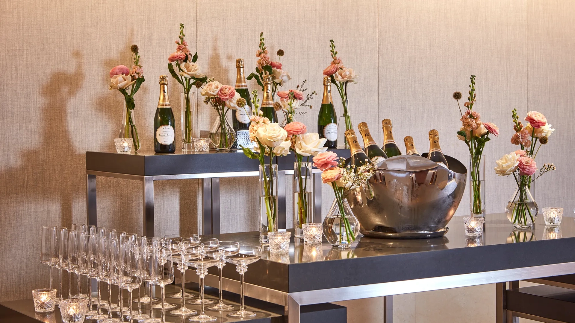 Champagne bar setup with bottles on ice, flutes, and vases of pastel flowers on modern black tables.