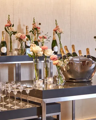 Champagne bar setup with bottles on ice, flutes, and vases of pastel flowers on modern black tables.