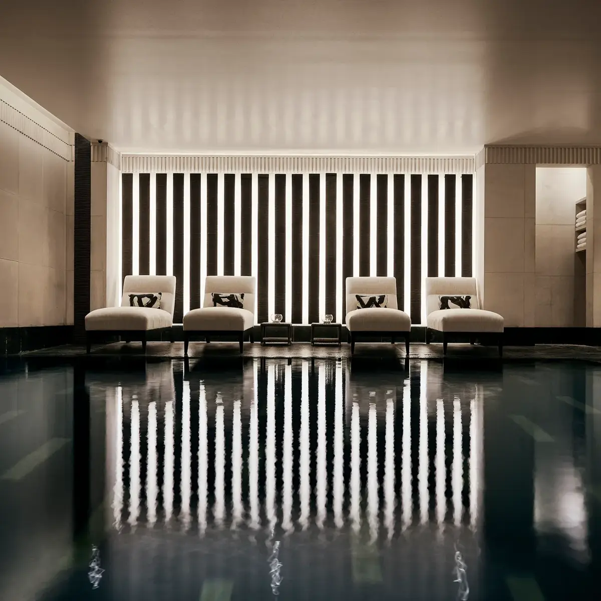 Indoor swimming pool with a sleek, contemporary design featuring a row of white loungers with cushions along a wall of vertical light panels. The dim lighting and calm reflections on the water create a serene, spa-like atmosphere.