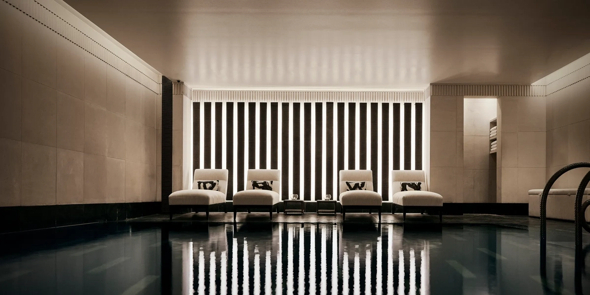 Indoor swimming pool with a sleek, contemporary design featuring a row of white loungers with cushions along a wall of vertical light panels. The dim lighting and calm reflections on the water create a serene, spa-like atmosphere.