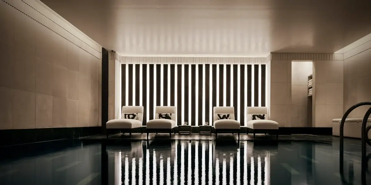 Indoor swimming pool with a sleek, contemporary design featuring a row of white loungers with cushions along a wall of vertical light panels. The dim lighting and calm reflections on the water create a serene, spa-like atmosphere.