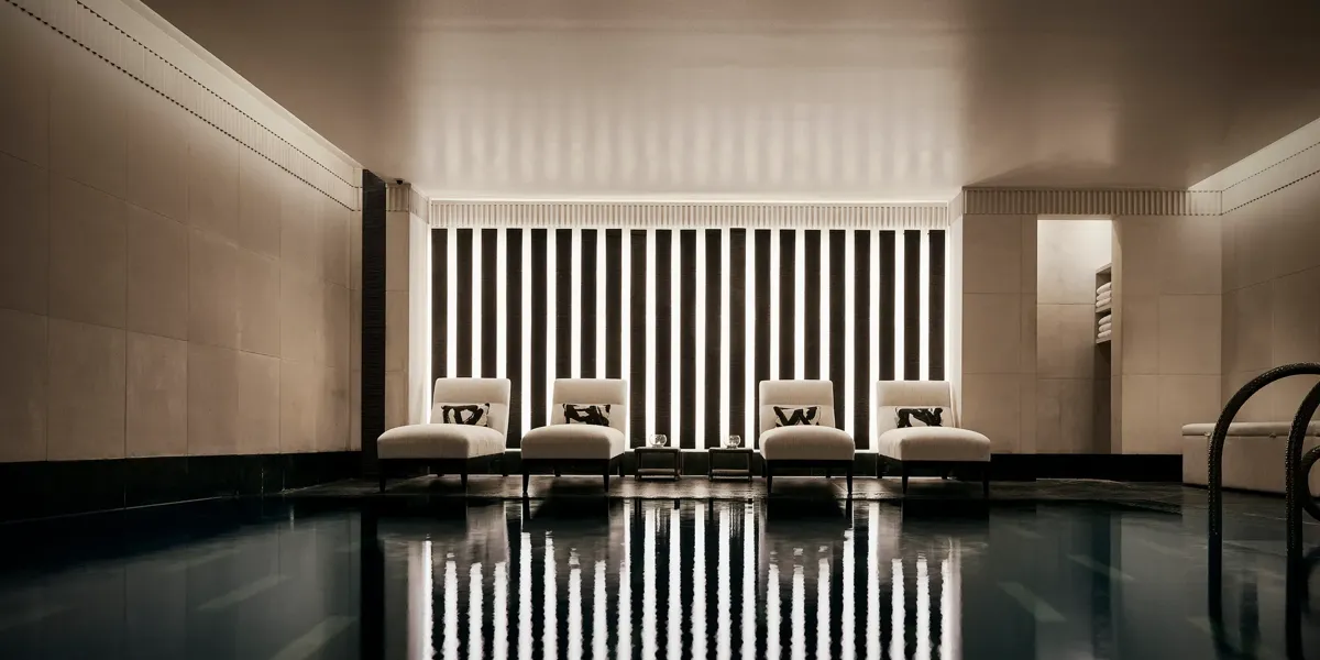 Indoor swimming pool with a sleek, contemporary design featuring a row of white loungers with cushions along a wall of vertical light panels. The dim lighting and calm reflections on the water create a serene, spa-like atmosphere.