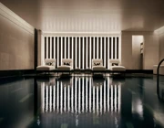 Indoor swimming pool with a sleek, contemporary design featuring a row of white loungers with cushions along a wall of vertical light panels. The dim lighting and calm reflections on the water create a serene, spa-like atmosphere.