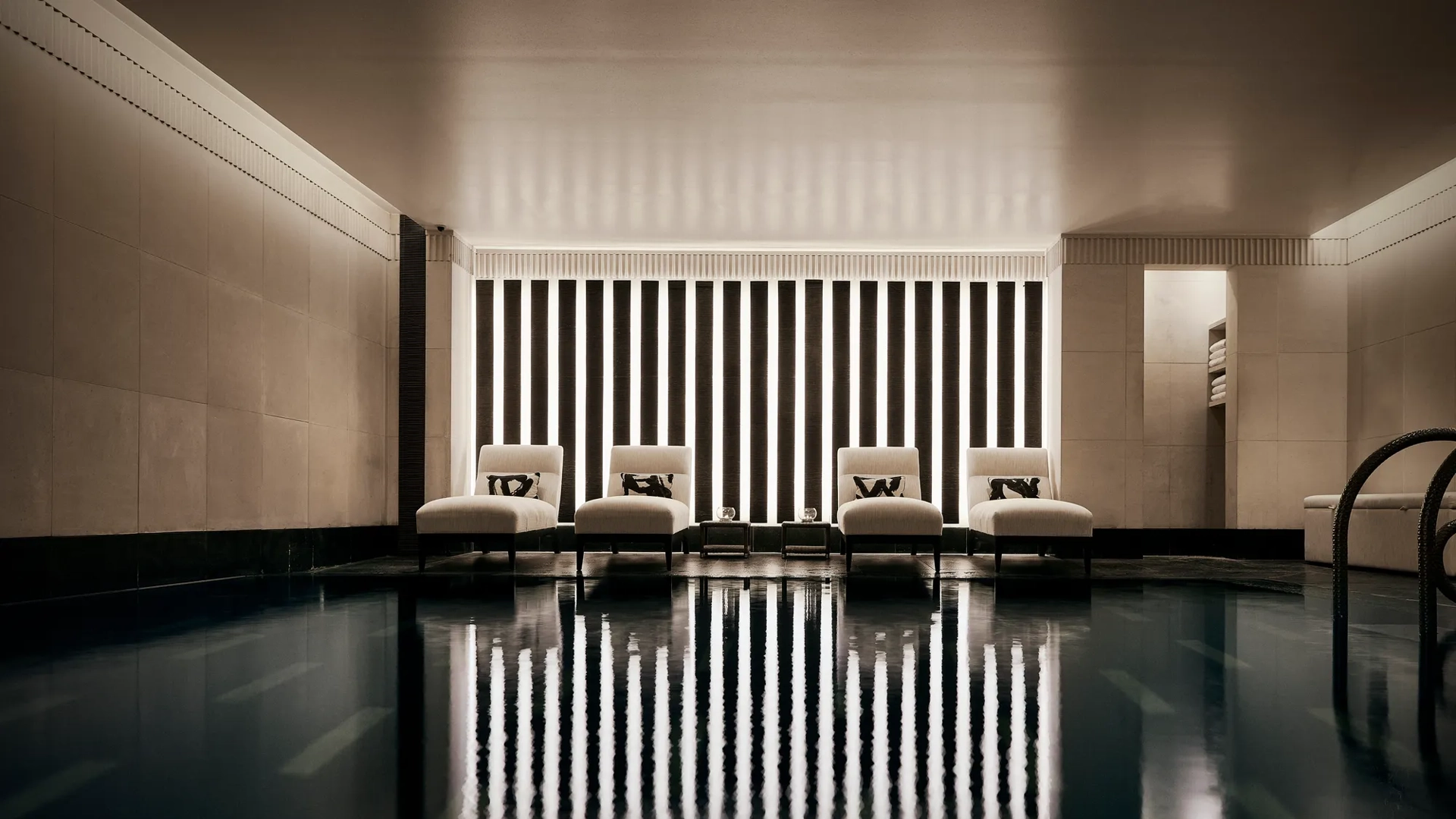 Indoor swimming pool with a sleek, contemporary design featuring a row of white loungers with cushions along a wall of vertical light panels. The dim lighting and calm reflections on the water create a serene, spa-like atmosphere.