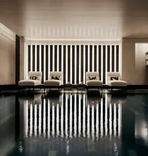 Indoor swimming pool with a sleek, contemporary design featuring a row of white loungers with cushions along a wall of vertical light panels. The dim lighting and calm reflections on the water create a serene, spa-like atmosphere.