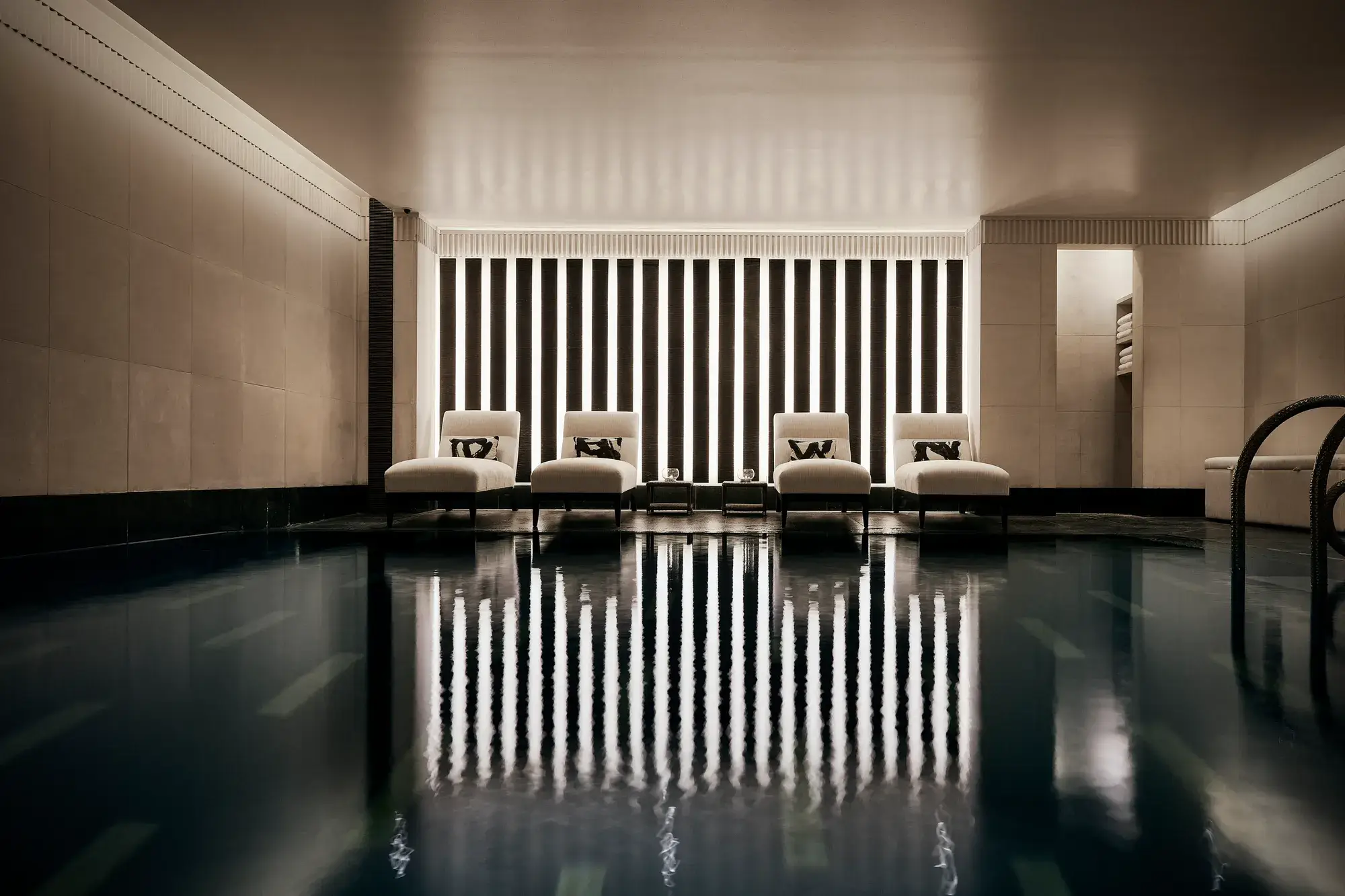 Indoor swimming pool with a sleek, contemporary design featuring a row of white loungers with cushions along a wall of vertical light panels. The dim lighting and calm reflections on the water create a serene, spa-like atmosphere.