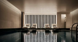 Indoor swimming pool with a sleek, contemporary design featuring a row of white loungers with cushions along a wall of vertical light panels. The dim lighting and calm reflections on the water create a serene, spa-like atmosphere.
