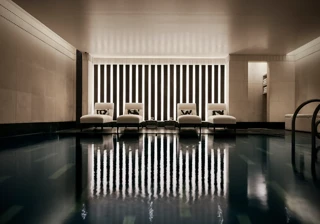 Indoor swimming pool with a sleek, contemporary design featuring a row of white loungers with cushions along a wall of vertical light panels. The dim lighting and calm reflections on the water create a serene, spa-like atmosphere.