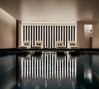 Indoor swimming pool with a sleek, contemporary design featuring a row of white loungers with cushions along a wall of vertical light panels. The dim lighting and calm reflections on the water create a serene, spa-like atmosphere.