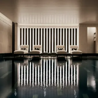 Indoor swimming pool with a sleek, contemporary design featuring a row of white loungers with cushions along a wall of vertical light panels. The dim lighting and calm reflections on the water create a serene, spa-like atmosphere.