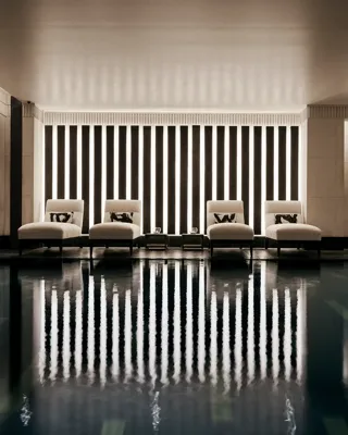 Indoor swimming pool with a sleek, contemporary design featuring a row of white loungers with cushions along a wall of vertical light panels. The dim lighting and calm reflections on the water create a serene, spa-like atmosphere.