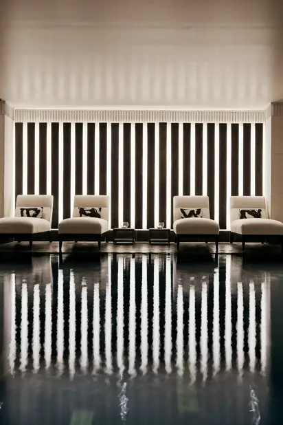 Indoor swimming pool with a sleek, contemporary design featuring a row of white loungers with cushions along a wall of vertical light panels. The dim lighting and calm reflections on the water create a serene, spa-like atmosphere.