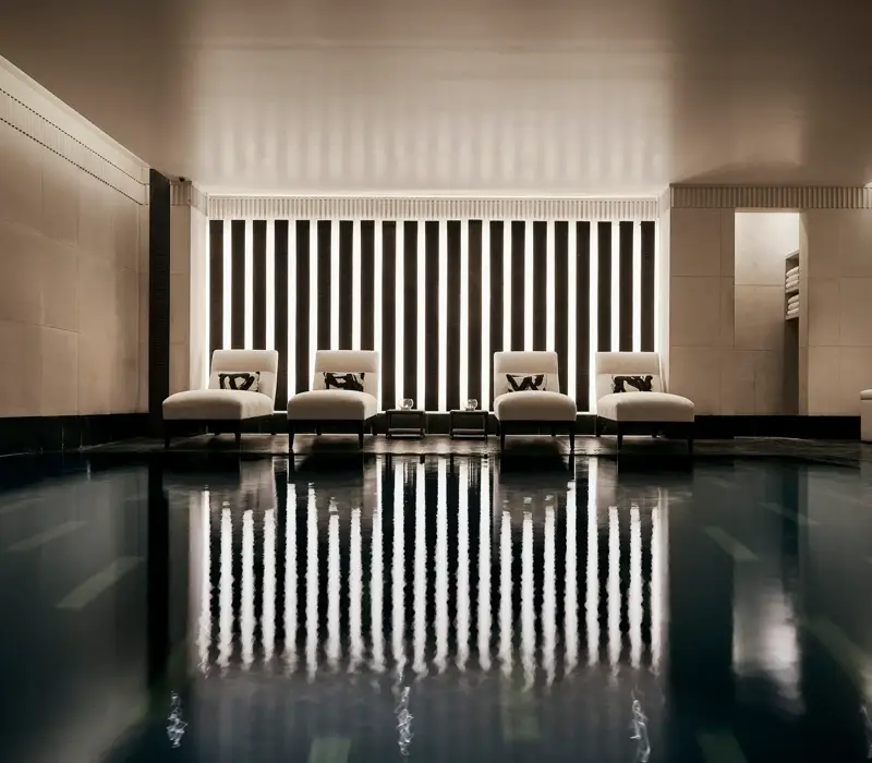 Indoor swimming pool with a sleek, contemporary design featuring a row of white loungers with cushions along a wall of vertical light panels. The dim lighting and calm reflections on the water create a serene, spa-like atmosphere.