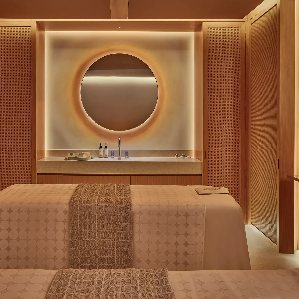 Warm-lit spa treatment room with two massage tables, textured throws, and a circular mirror above a stone countertop.