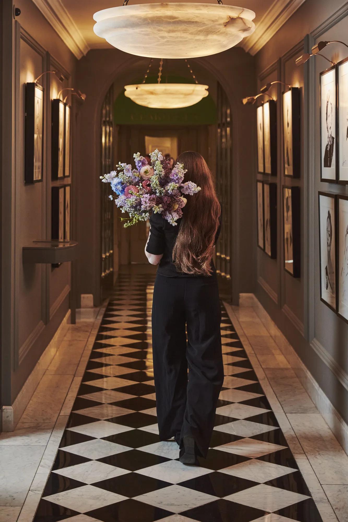 Person walking through an elegant hallway holding a large bouquet, with framed artwork, soft lighting and a patterned floor.