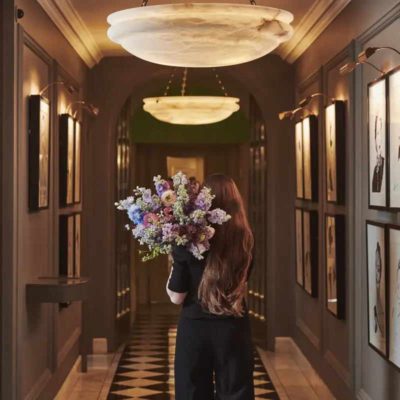Person walking through an elegant hallway holding a large bouquet, with framed artwork, soft lighting and a patterned floor.