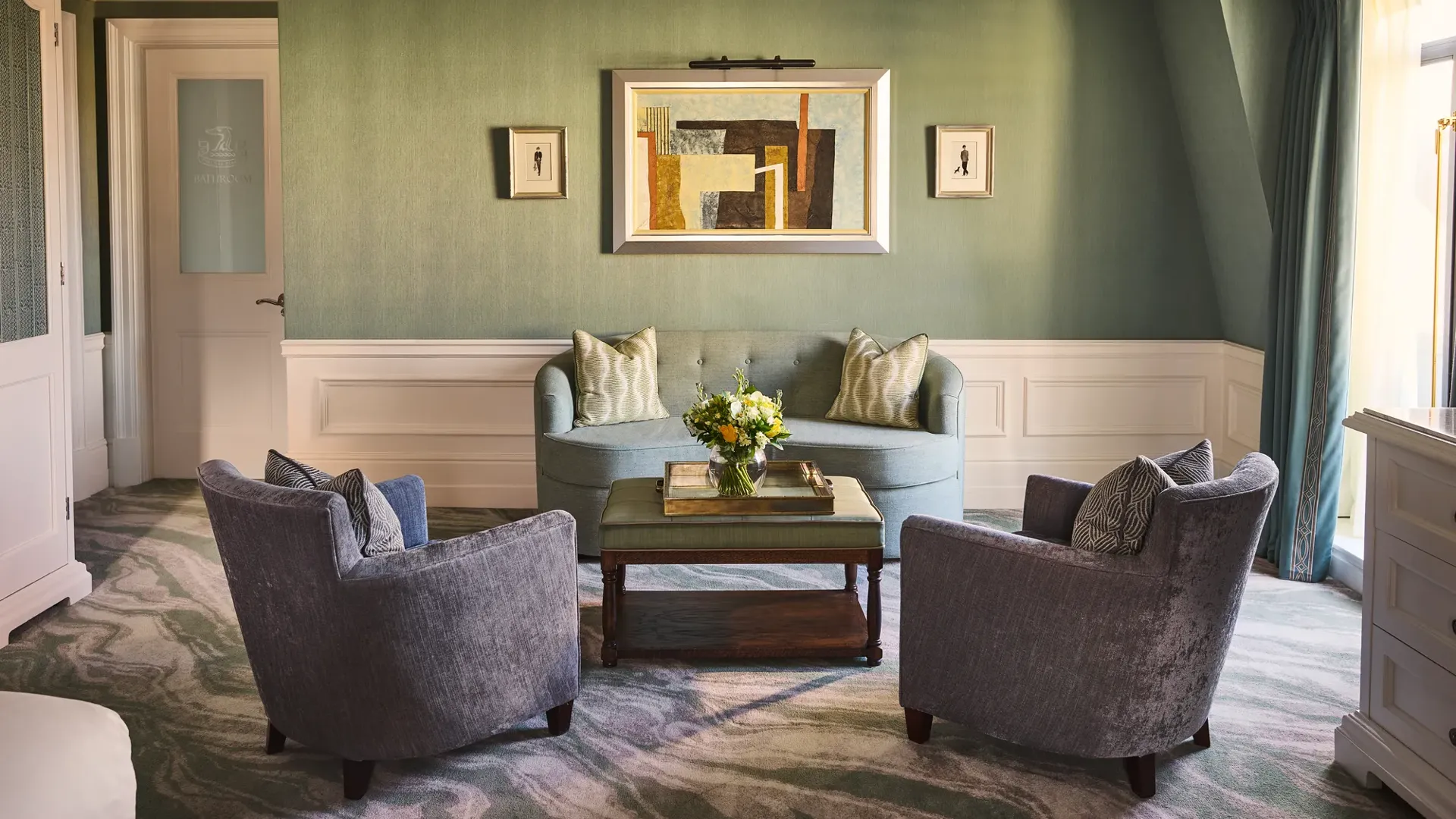 Elegant sitting area with green walls, blue sofa, two armchairs, coffee table with flowers and framed artwork above.
