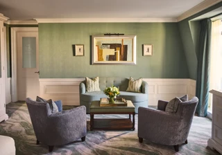 Elegant sitting area with green walls, blue sofa, two armchairs, coffee table with flowers and framed artwork above.