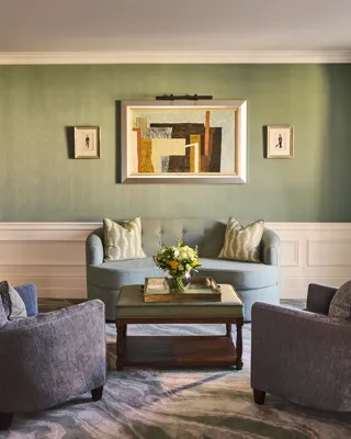 Elegant sitting area with green walls, blue sofa, two armchairs, coffee table with flowers and framed artwork above.