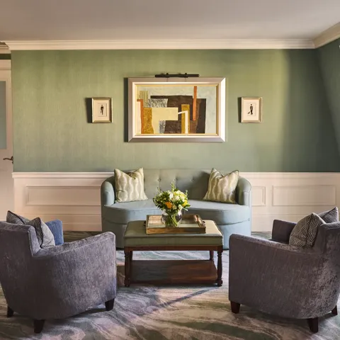 Elegant sitting area with green walls, blue sofa, two armchairs, coffee table with flowers and framed artwork above.
