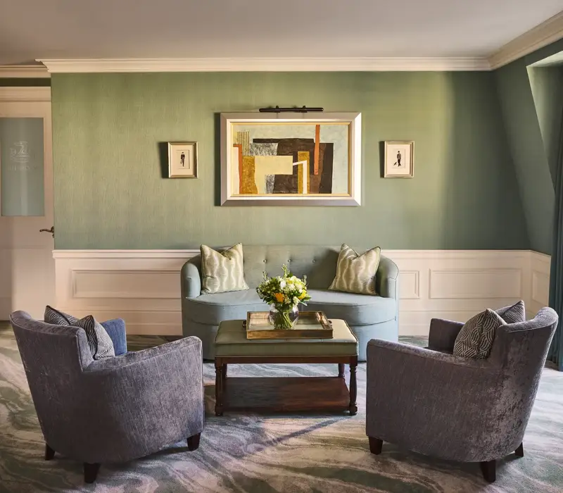 Elegant sitting area with green walls, blue sofa, two armchairs, coffee table with flowers and framed artwork above.