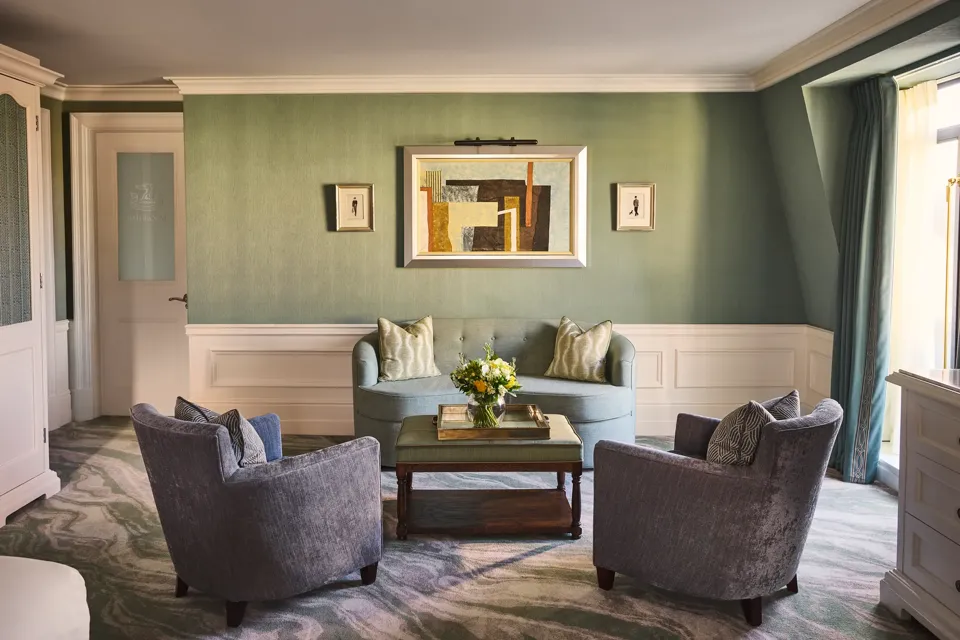 Elegant sitting area with green walls, blue sofa, two armchairs, coffee table with flowers and framed artwork above.