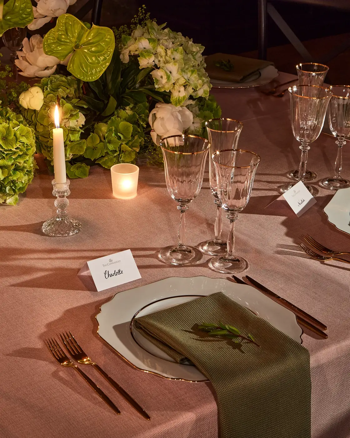 Candlelit table setting with elegant glassware, white plates and green napkins, surrounded by lush green and white floral arrangements.
