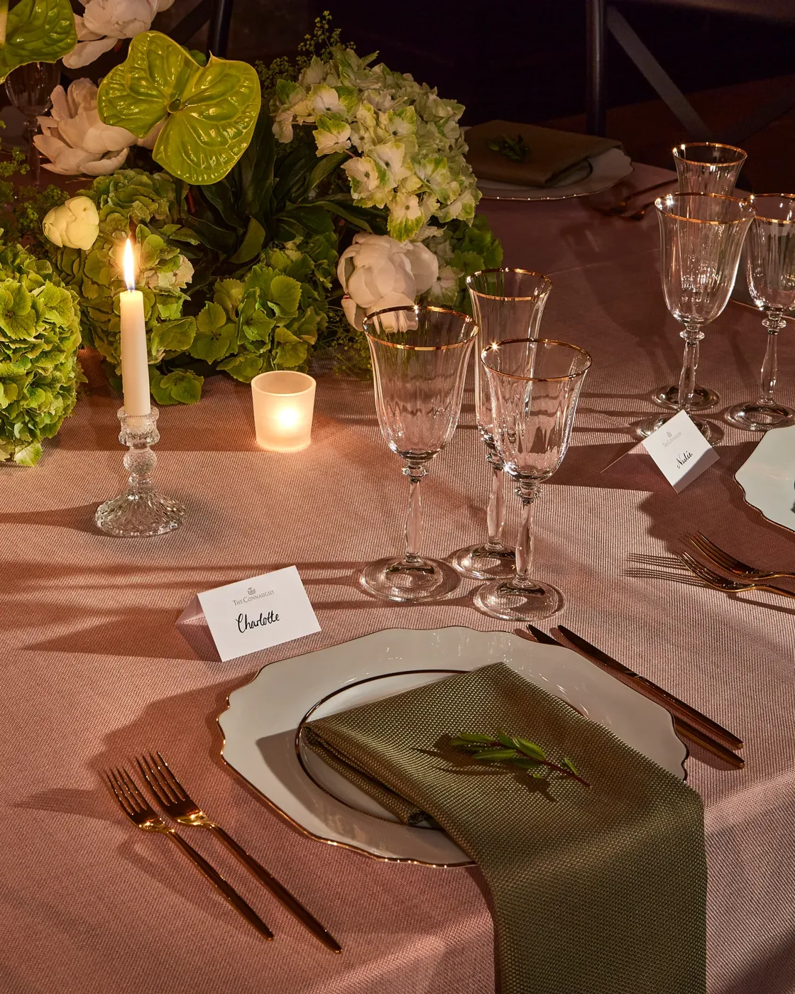 Candlelit table setting with elegant glassware, white plates and green napkins, surrounded by lush green and white floral arrangements.