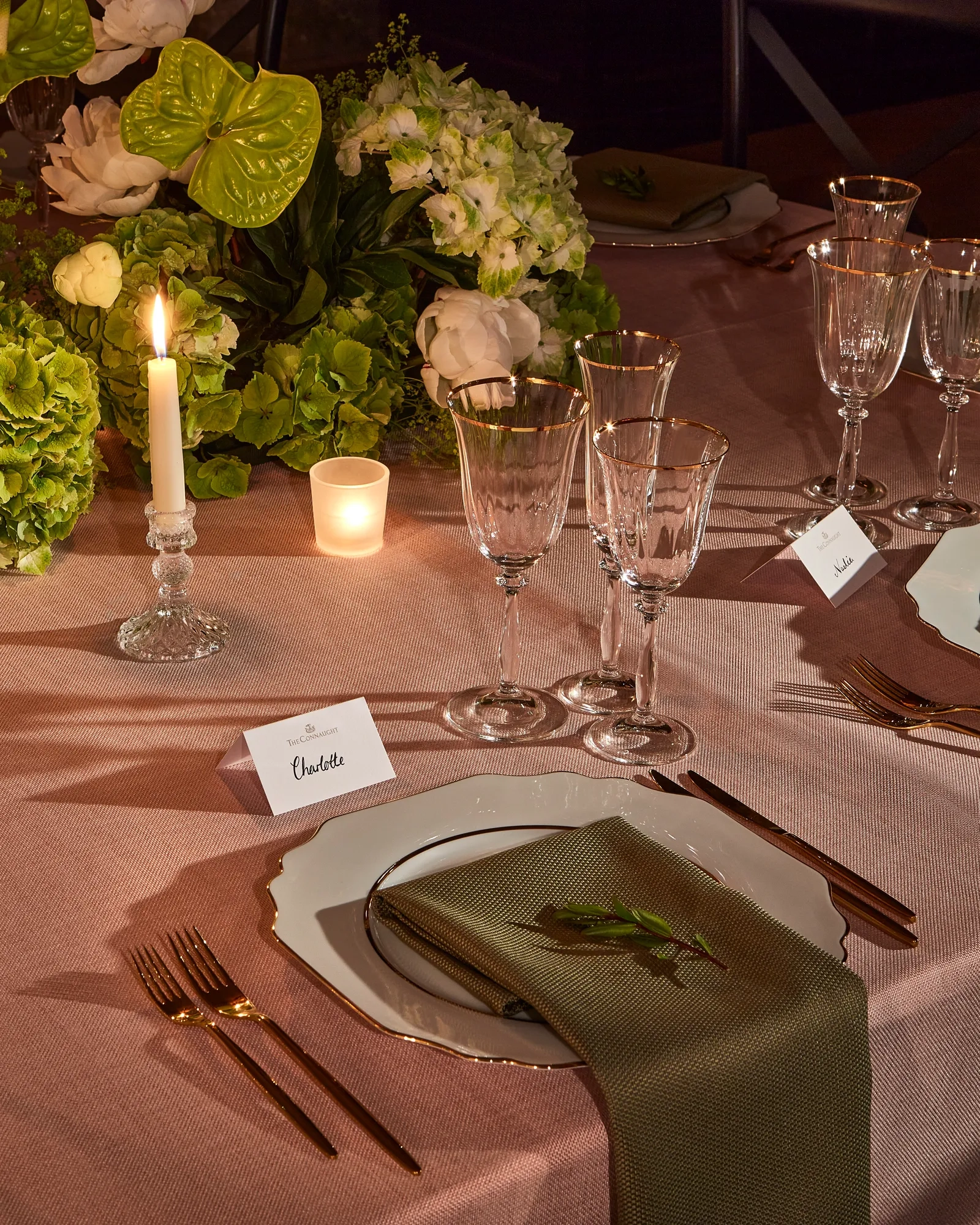 Candlelit table setting with elegant glassware, white plates and green napkins, surrounded by lush green and white floral arrangements.