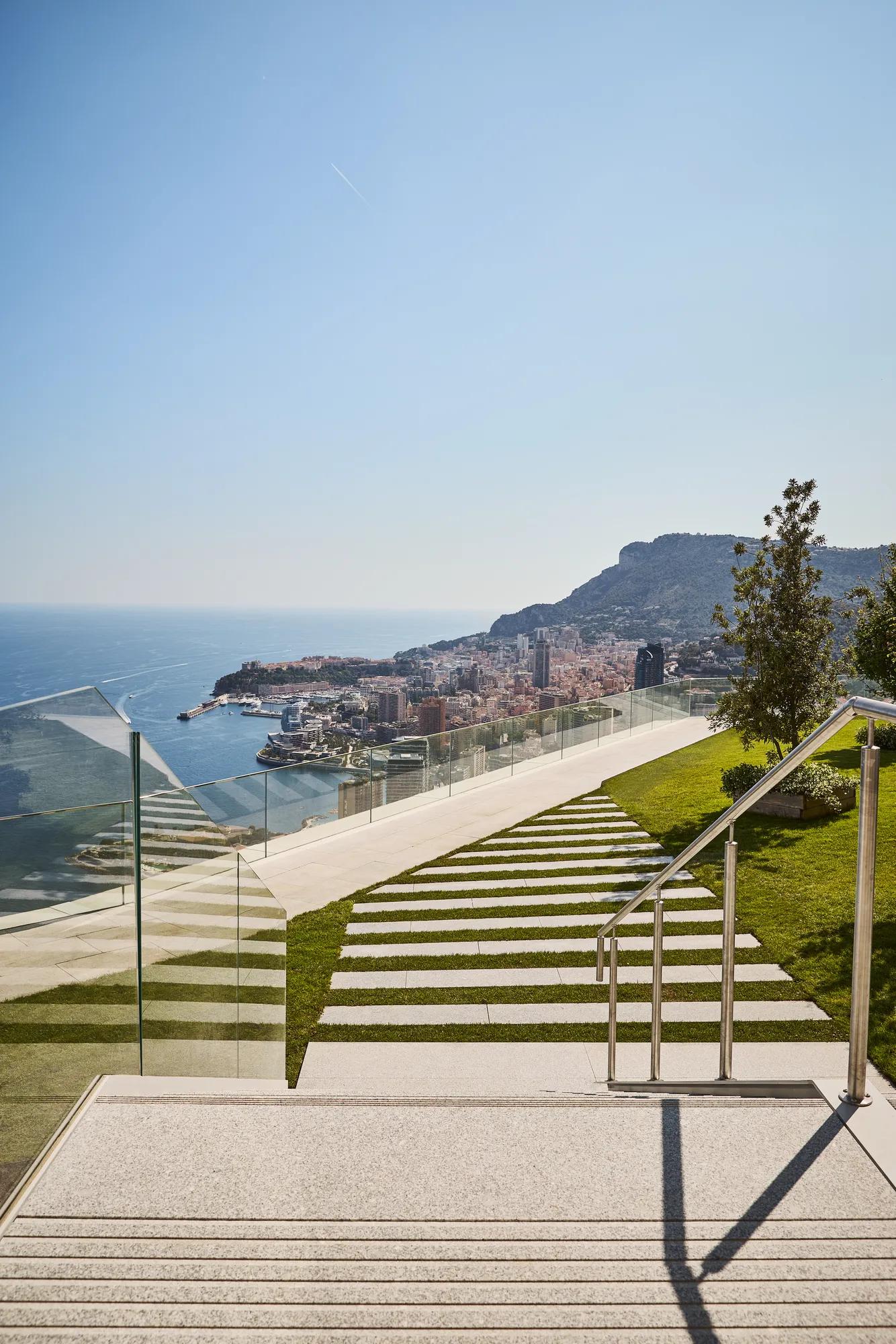 Modern outdoor steps with striped stone and grass design, bordered by glass railings, leading to a terrace with panoramic views of the sea, city, and hills.