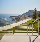 Modern outdoor steps with striped stone and grass design, bordered by glass railings, leading to a terrace with panoramic views of the sea, city, and hills.