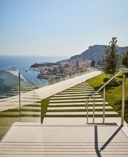 Modern outdoor steps with striped stone and grass design, bordered by glass railings, leading to a terrace with panoramic views of the sea, city, and hills.