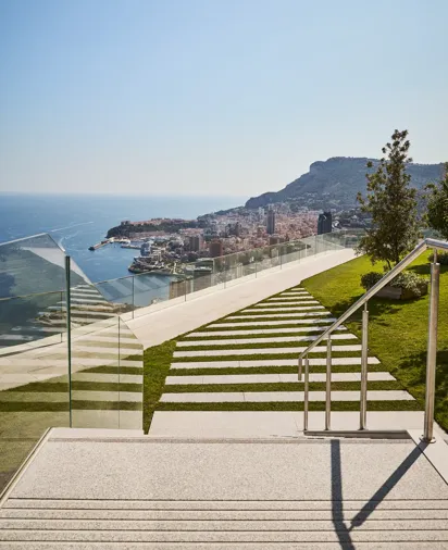 Modern outdoor steps with striped stone and grass design, bordered by glass railings, leading to a terrace with panoramic views of the sea, city, and hills.