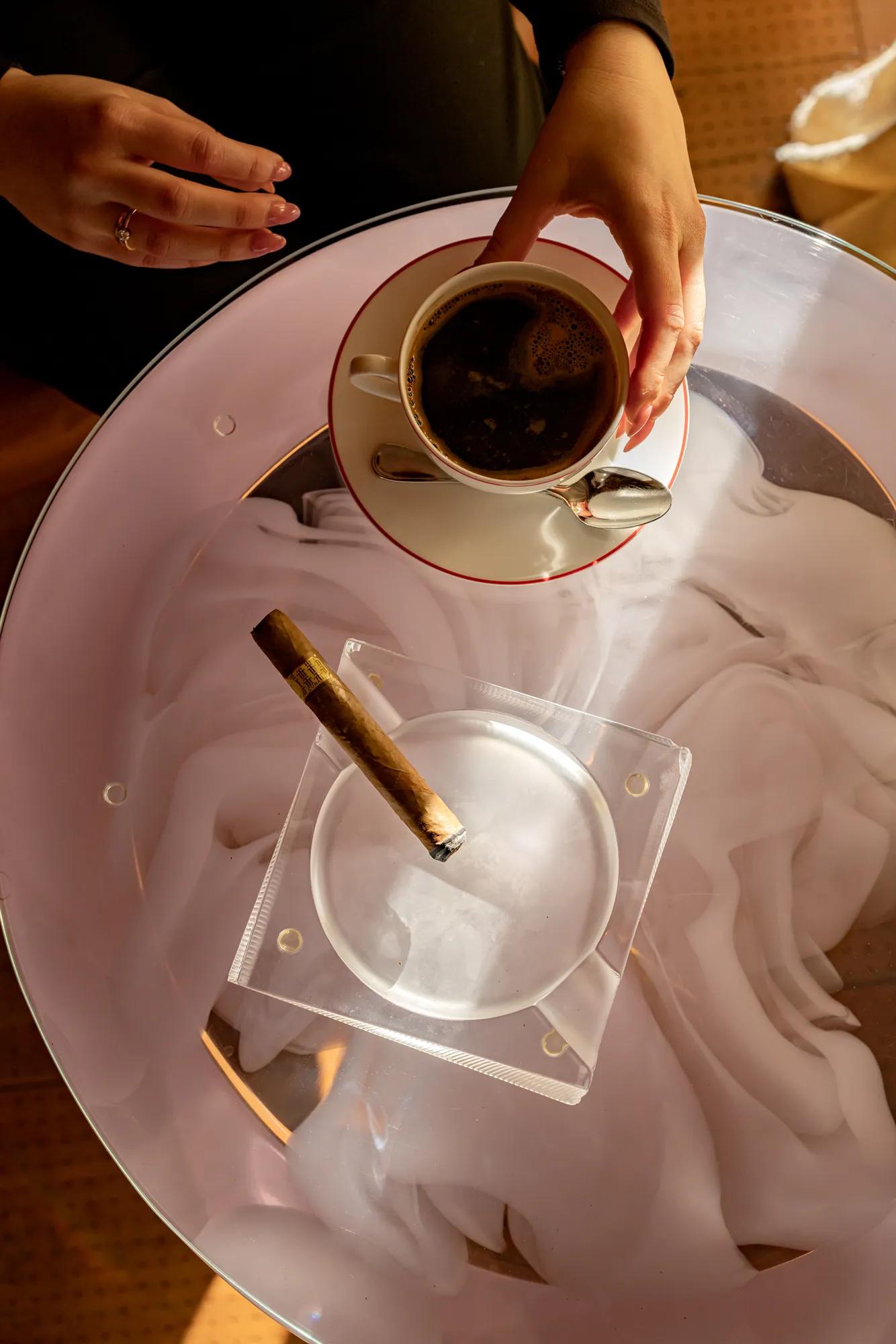 Hands holding a cup of coffee above a glass table with a cigar resting in an ashtray