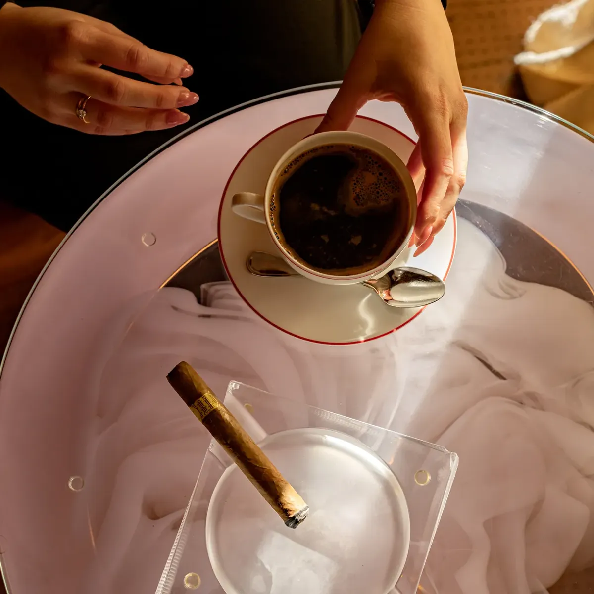 Hands holding a cup of coffee above a glass table with a cigar resting in an ashtray