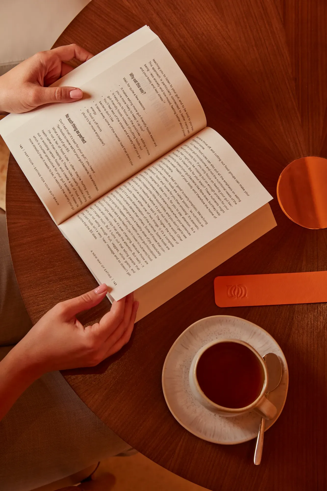 Hands holding an open book on a round wooden table with a coffee cup and leather bookmark nearby.