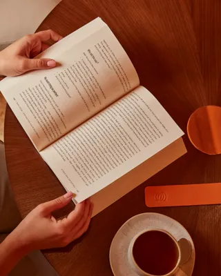 Hands holding an open book on a round wooden table with a coffee cup and leather bookmark nearby.