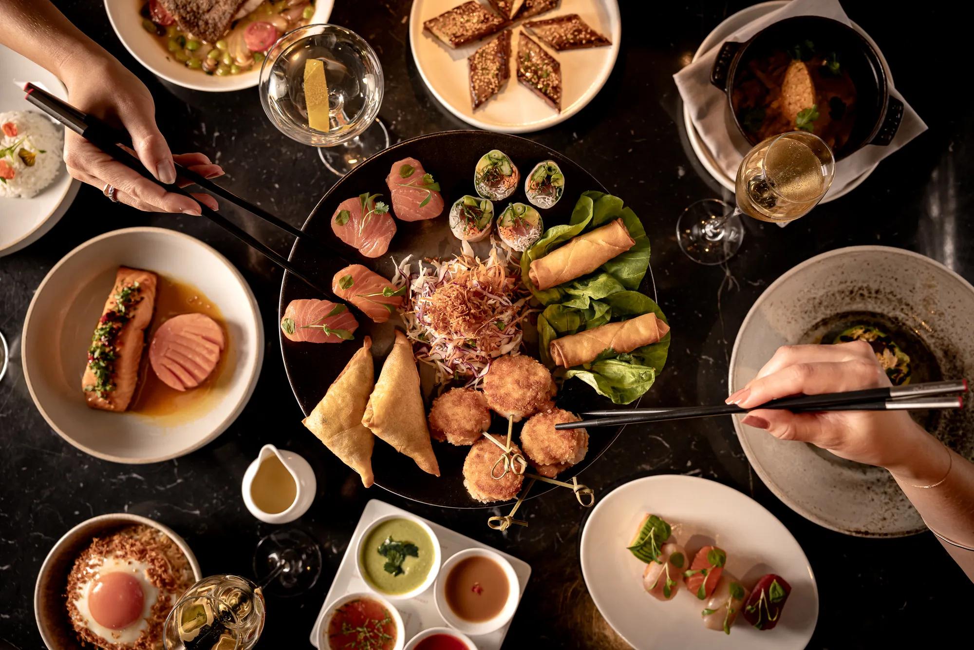 Overhead view of a table filled with sharing plates, sushi, spring rolls and drinks, with guests serving food using chopsticks.