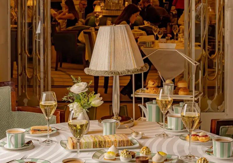 Refined afternoon tea setting with striped china, pastries, and champagne glasses arranged on a round white tablecloth. A softly lit lamp and floral arrangement sit at the center, with guests enjoying tea service in the elegant room beyond.